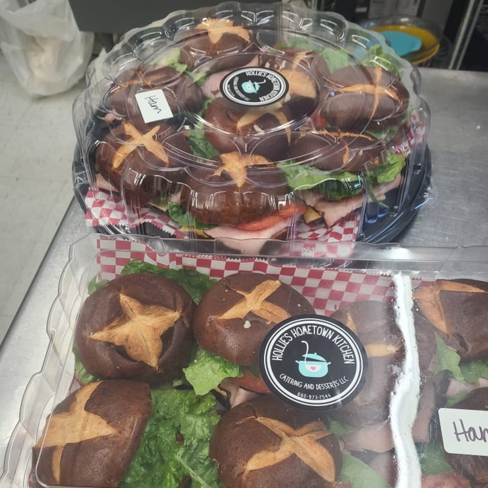 Two catering trays of Hollie's Hometown Kitchen ham sandwiches on pretzel buns with lettuce and tomato.
