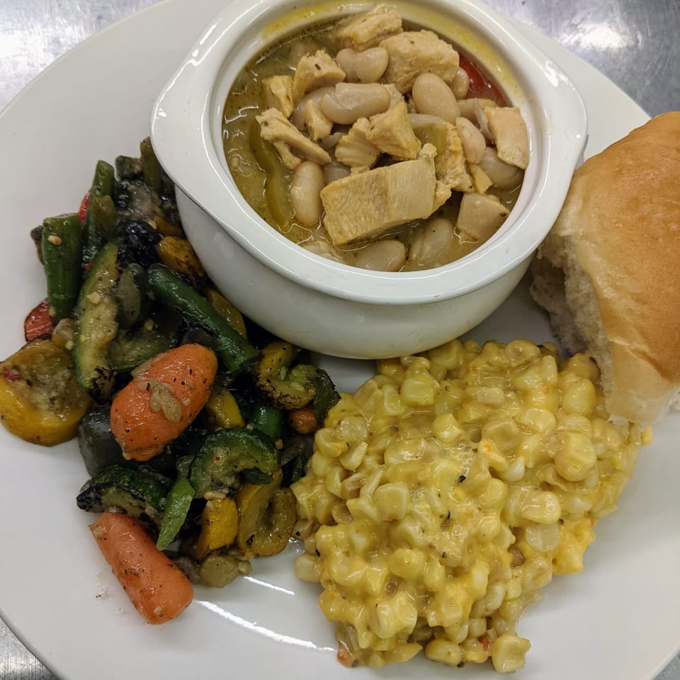 A white bowl of chicken and white bean chili, with roasted vegetables, creamed corn, and a roll.