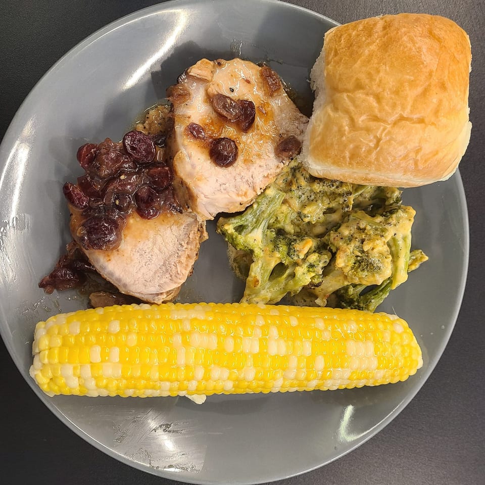 Pork chops with raisin sauce, cheesy broccoli, corn on the cob, and a dinner roll on a gray plate.