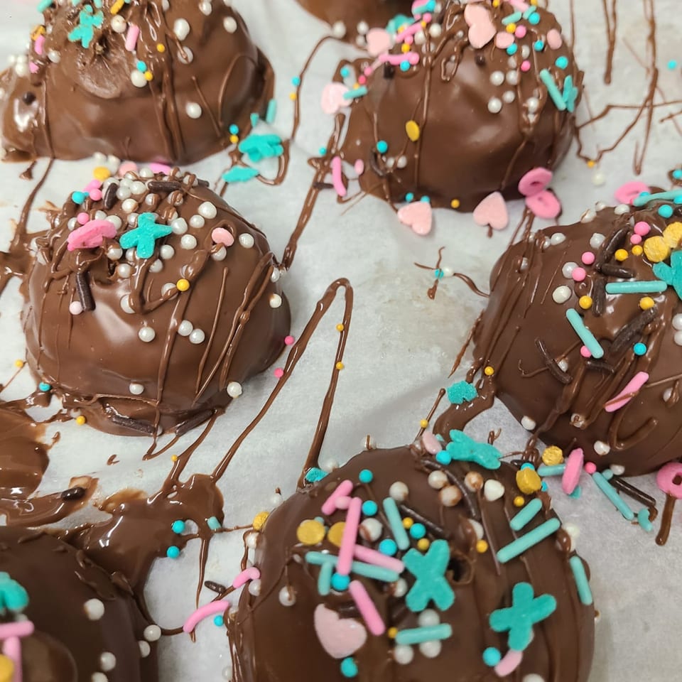 Several chocolate-covered desserts with colorful sprinkles and drizzles on white parchment paper.